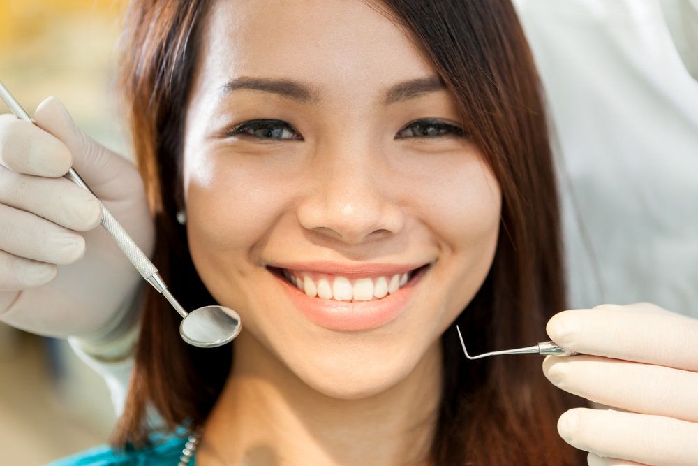 <p>A closeup of a young woman's face with a dentist holding a mirror and dental pick in front of her.</p>